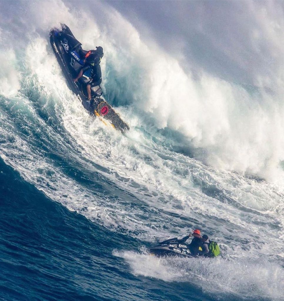 Picure of the Hawaii water patrol on a jet ski going over a massive wave wearing the orange shelta sun hat