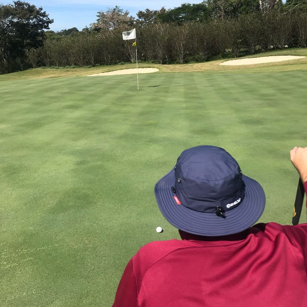 View from behind a golfer looking at a pin on the green of a golf course wearing a shelta hat